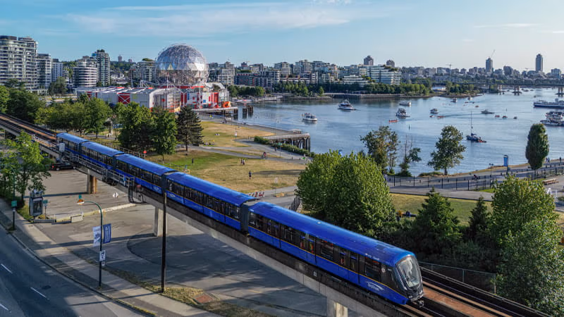 New Mark V train travels along the waterfront with a city skyline in the background.