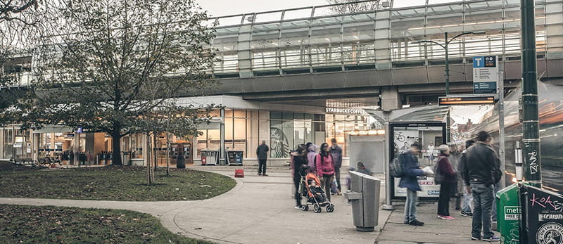 People lining up at a bus stop in front of a SkyTrain station and retail shops