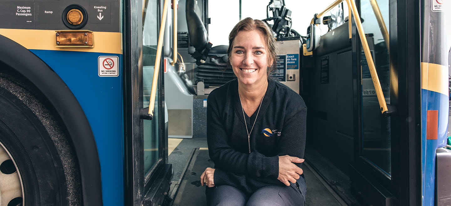 A woman sitting outside a conventional bus. She's crossing her arms and smiling softly at the camera