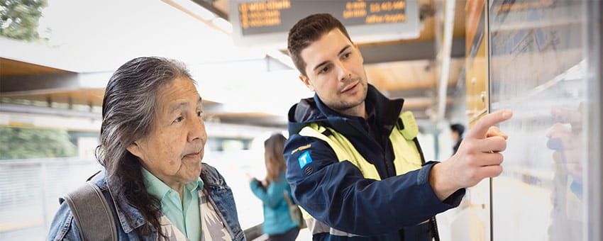 A BCRTC SkyTrain Attendant wearing a TransLink uniform is assisting a customer at a transit station.