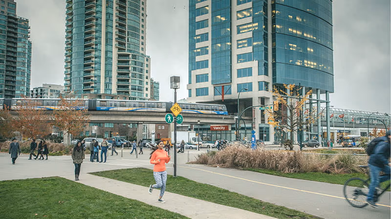 A cyclist, a runner, and a group of people walking on a sidewalk next to tall buildings