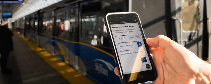 Mobile phone on ‘Transit Alerts’ page held up in front of SkyTrain at station platform