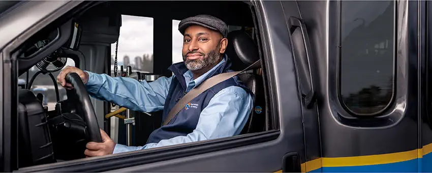A male Community Shuttle driver grasping the steering wheel with one hand and smiling directly at the camera