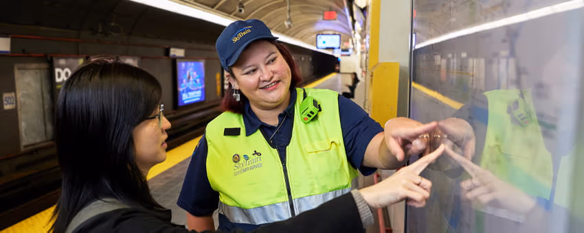 A SkyTrain Attendant in a safety vest and cap helps a passenger by pointing at a transit map.