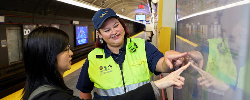A SkyTrain Attendant in a safety vest and cap helps a passenger by pointing at a transit map.