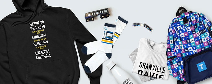 Merchandise available on the TransLink store including including a black hoodie with transit route names, socks with bus designs, a white water bottle, a backpack with a colorful transit logo pattern, a Granville T-shirt, and model buses.