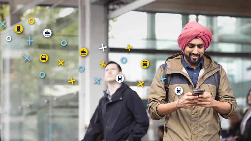 A passenger stands in front of a station, looking at their phone, with yellow, teal, and white bus, seabus, and skytrain icons floating above the image.