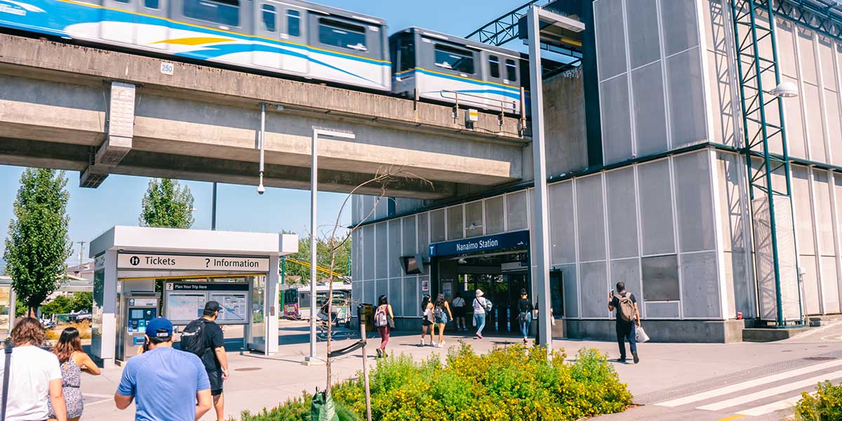 People walking toward Nanaimo Station entrance on a sunny day, with a train passing overhead on an elevated track