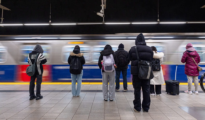 A group of bundled-up commuters waits on a brightly lit SkyTrain platform as a train speeds past.