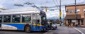 A TransLink trolleybus passing through an intersection, with overhead wires, traffic lights, and nearby shops visible in the background.