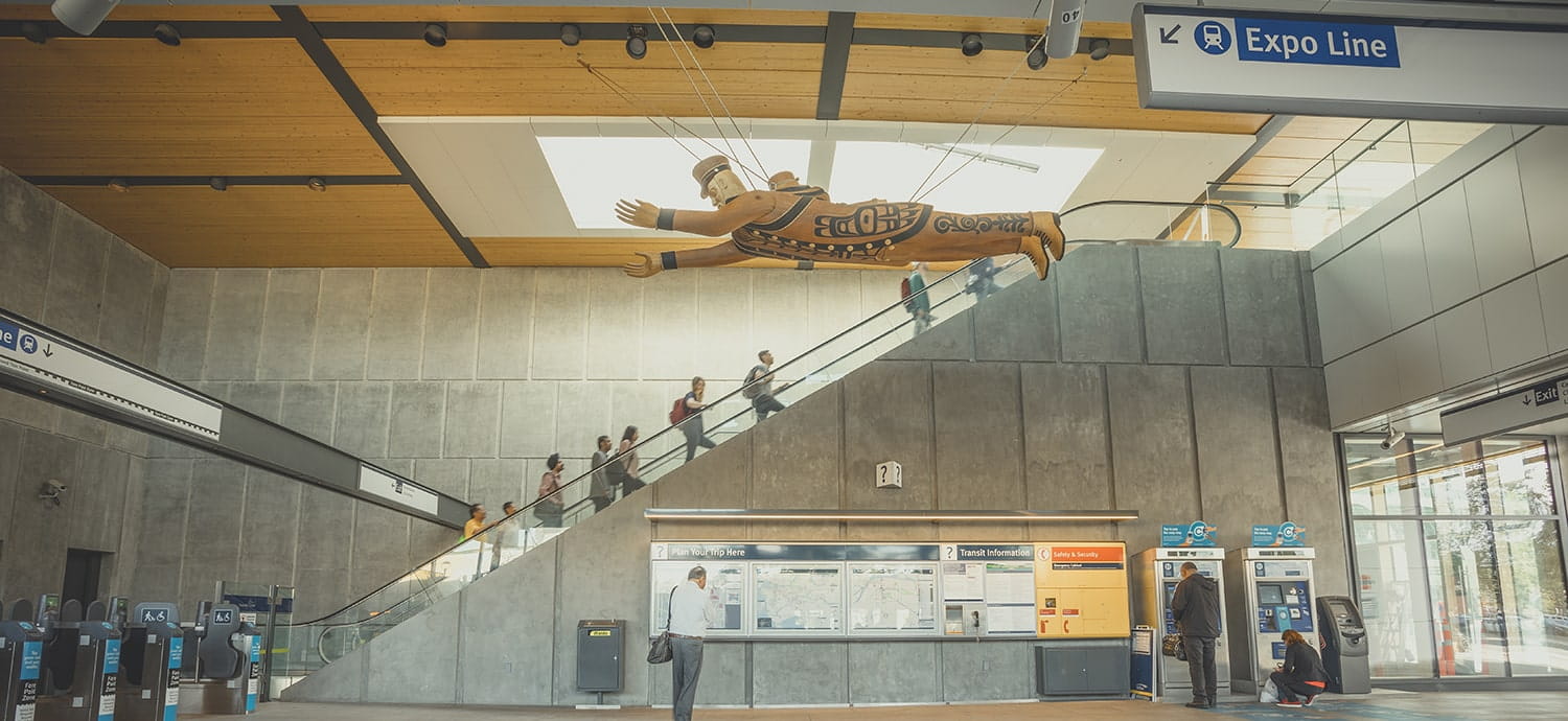The interior of a SkyTrain Station with people going up escalators and viewing the overhanding artwork