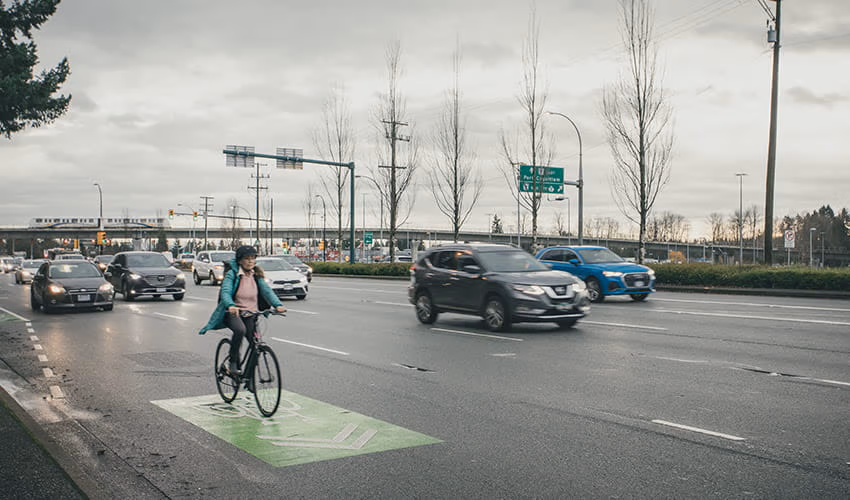 A women cyclist riding on a designated bike lane alongside moving traffic on a cloudy day, with cars and road signs visible in the background.