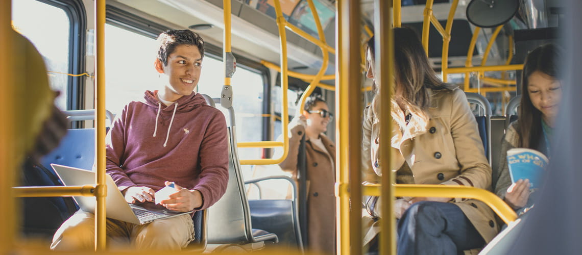 A young man with a laptop chatting with another passenger while riding the bus