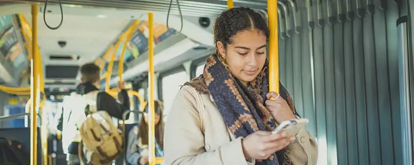 A young woman on the bus looking at her phone while standing and holding a stanchion