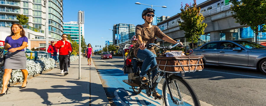 A woman smiling while riding her bike down a designated blue bike lane with pedestrians on the sidewalk to her left