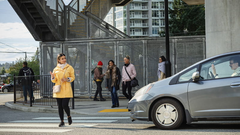 A woman in a yellow coat crosses at a crosswalk as a car waits and pedestrians walk behind her.
