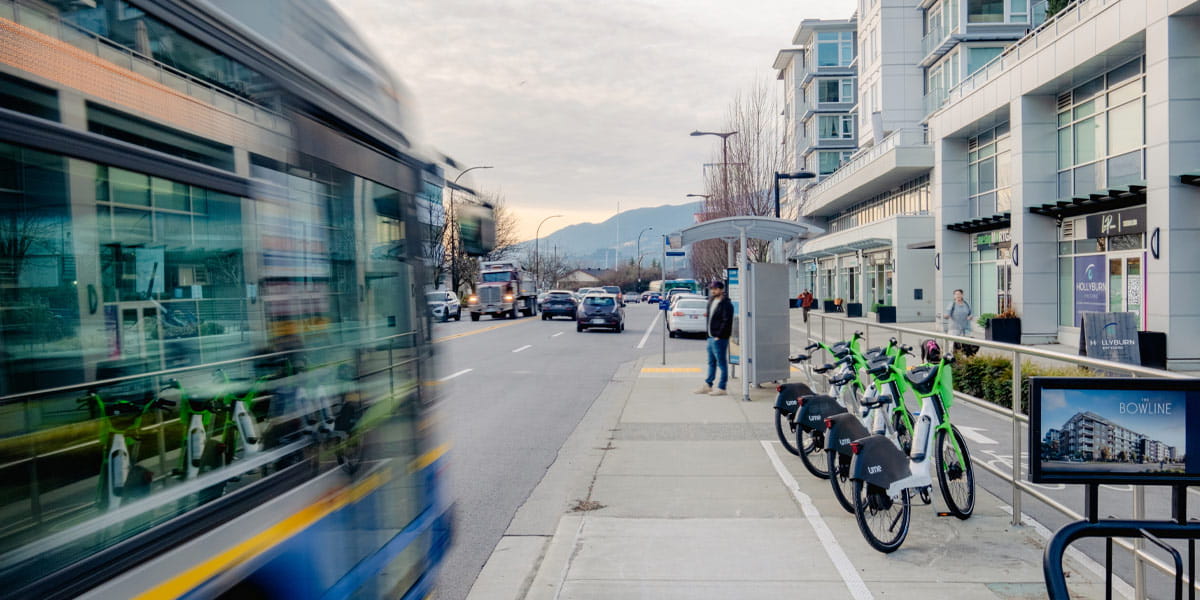 City street with a moving bus, people at a bus stop, and parked shared e-bikes near modern buildings.