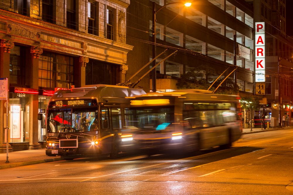 Two buses, including NightBus travelling on downtown Vancouver street at night.