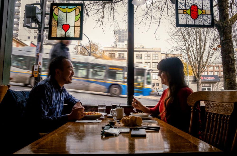 Man and woman sitting across from each other at restaurant booth near window, with passerby looking in and bus passing by.
