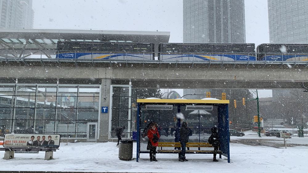 Snow falling at Metrotown SkyTrain Station with SkyTrain passing by overhead and people waiting under bus shelter.