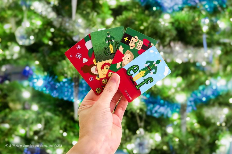 Hand holding four Elf-themed Compass Cards in front of Christmas tree.
