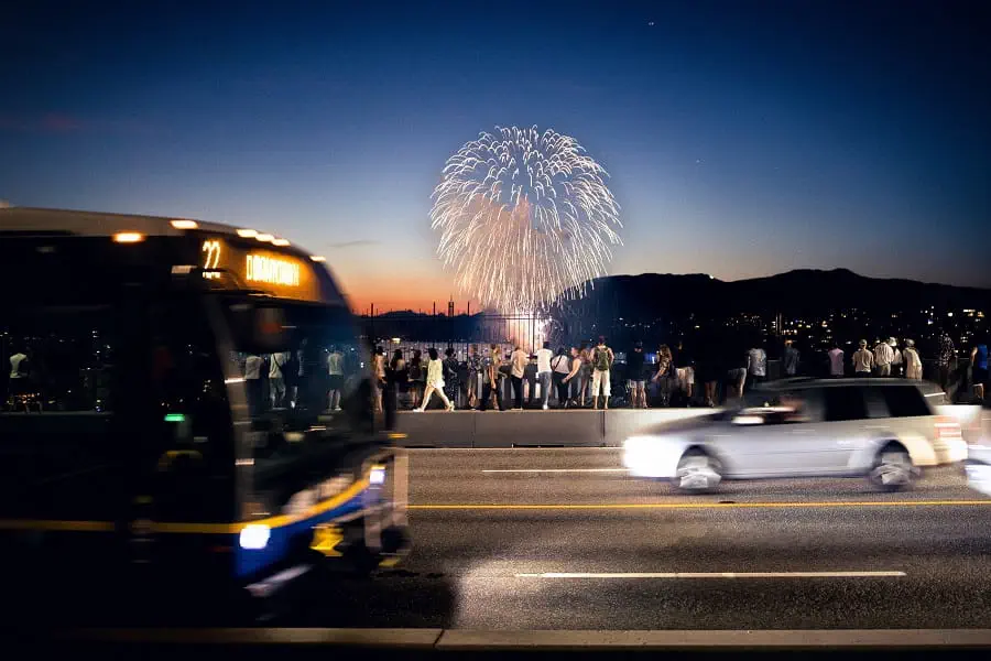 Bus and cars passing fireworks spectators on bridge.
