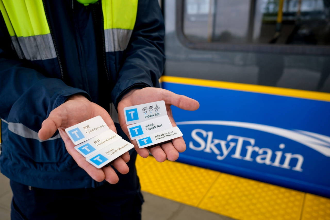 A SkyTrain Attendant, whose face is not visible, is holding six language pins in their hands. Each pin displays a different language, including one that displays the ability to speak ASL or American Sign Language
