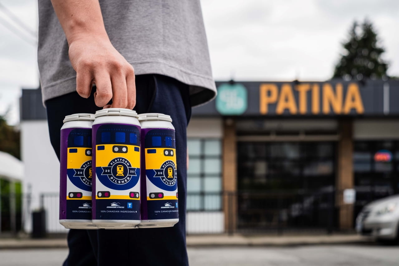 A man is carrying a four-pack of Platform Pilsner, walking along the street in front of the Patina Brewery