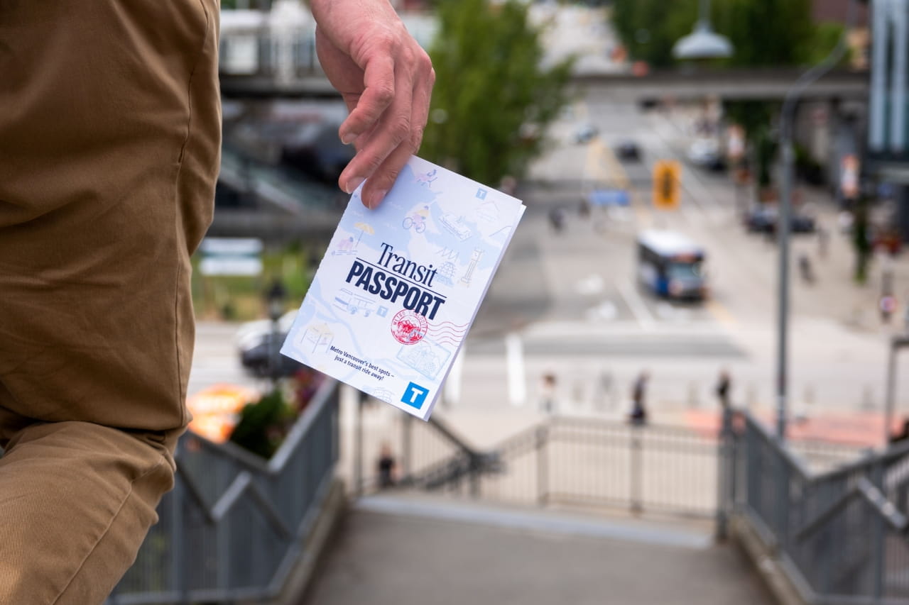a hand is holding a copy of the Transit Passport. The person is walking down a flight of stairs with a busy beautiful summer day outside.