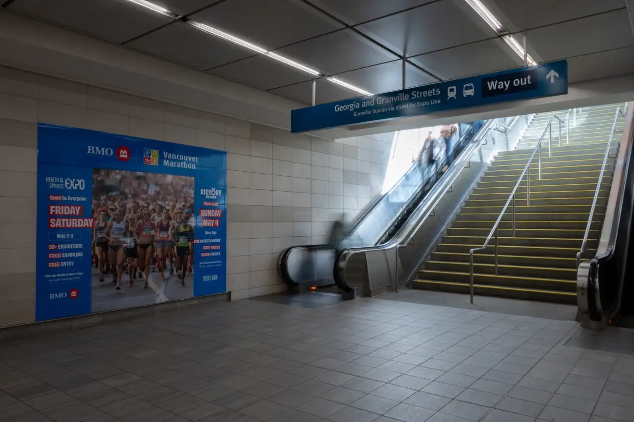 A large poster advertisement for the 2025 BMO Vancouver Marathon is on display at the Vancouver-City Centre Station concourse on the Canada Line. There are stairs and escalators leading riders up to the intersection of West Georgia and Granville Street.