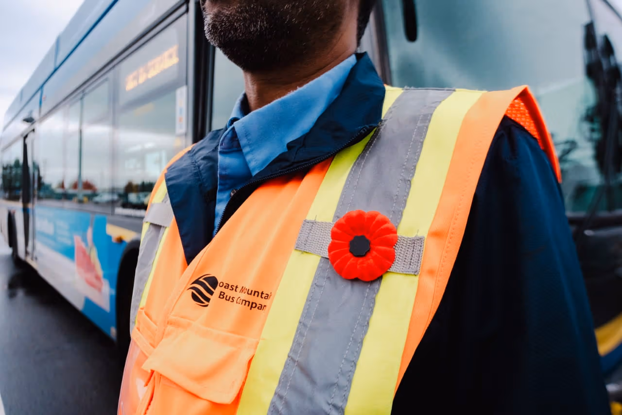 A bus operator is wearing a poppy on his chest, over his high-visibility safety vest. His work jacket reads Coast Mountain Bus Company.