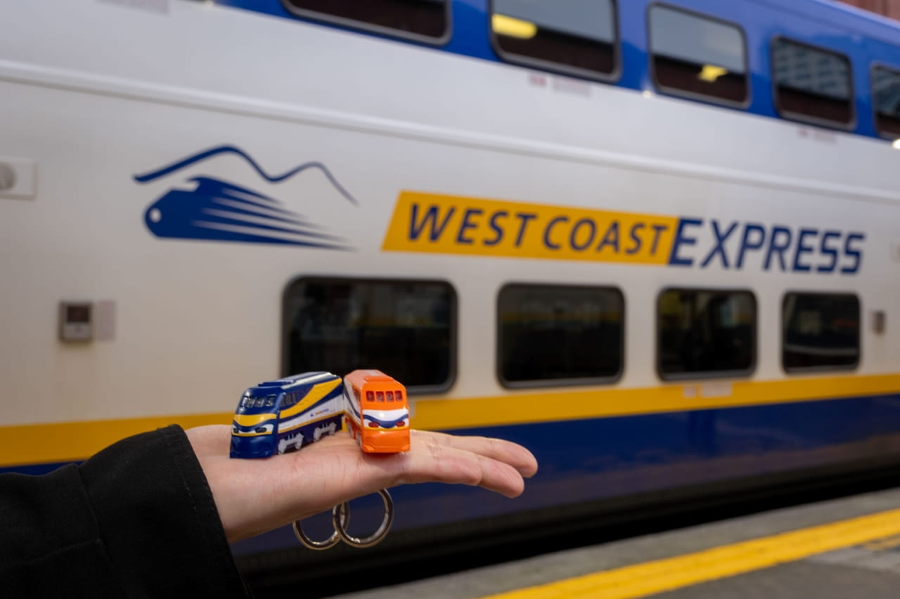 Two Compass Mini-West Coast Express are being held up in someone's hand, in front of a real West Coast Express train car. The two Compass Minis are different from one another. One is blue, indicating an Adult Compass product, and the other is orange, indicating a Concession Compass product.