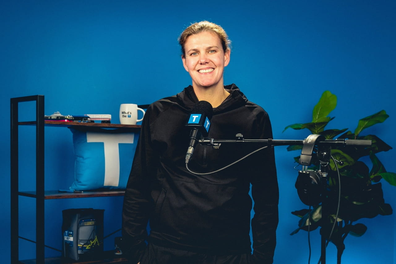 Christine Sinclair stands smiling in a podcast studio in front of a blue backdrop, speaking into a microphone with a TransLink logo. A shelf behind her holds TransLink-branded items, and a plant sits beside studio equipment