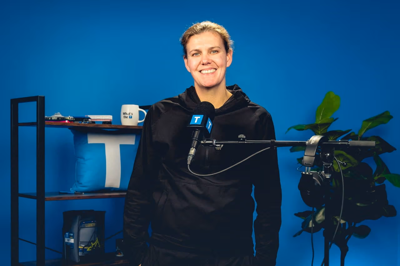 Christine Sinclair stands smiling in a podcast studio in front of a blue backdrop, speaking into a microphone with a TransLink logo. A shelf behind her holds TransLink-branded items, and a plant sits beside studio equipment