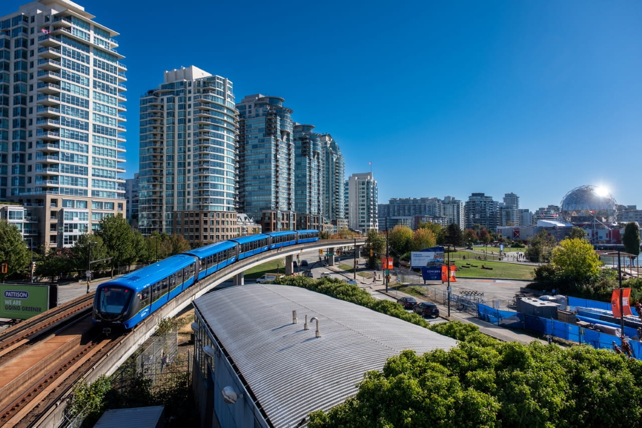 Elevated SkyTrain traveling along a curved track through a modern urban neighbourhood, with high-rise residential buildings on the left and a park and geodesic dome visible in the distance under a clear blue sky.