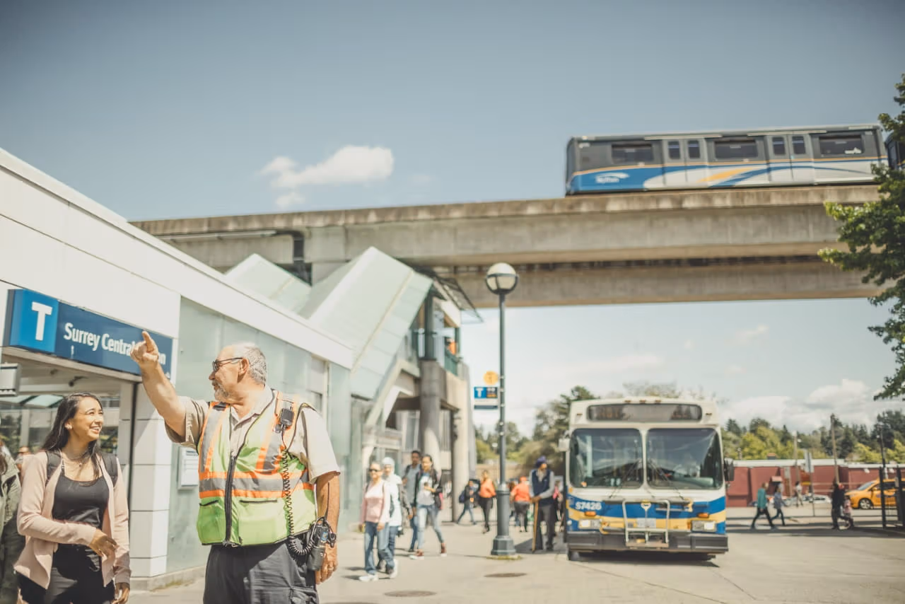Transit employee wearing a safety vest is helping to direct a customer in the right direction. They are standing just outside Surrey Central Station according to the sign behind them.
