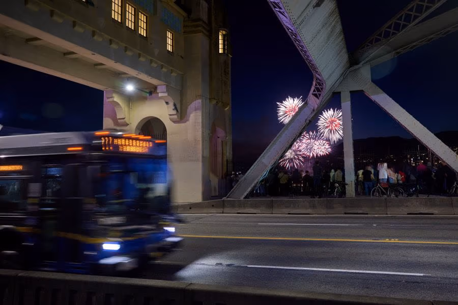 Bus driving over Burrard Street Bridge at night with crowd of people watching fireworks on bridge sidewalk