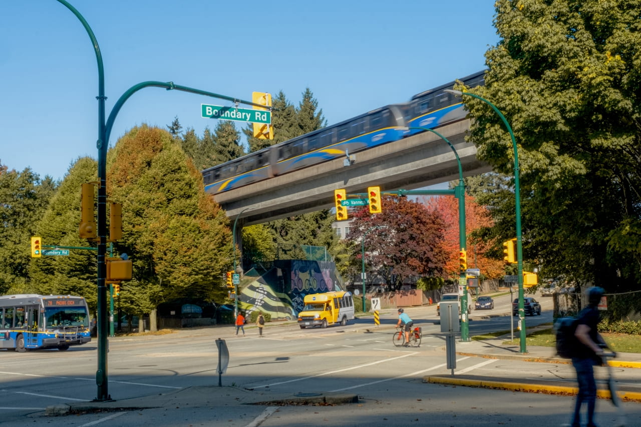 An elevated SkyTrain moves across a track above Boundary Road in Vancouver as buses, a cyclist, and pedestrians share a busy intersection surrounded by trees with fall colours.