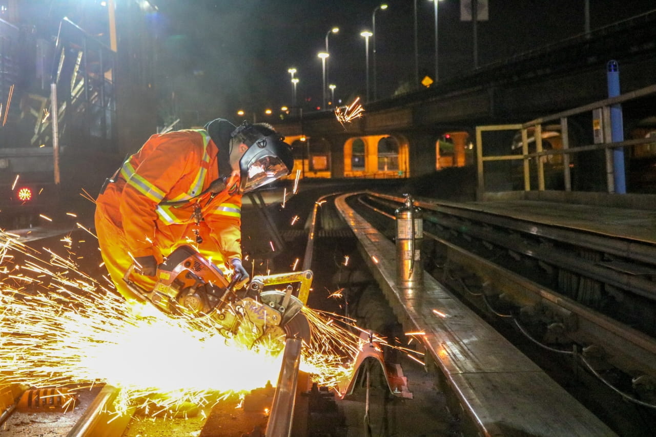 Maintenance worker in high-visibility orange protective gear uses a power saw to cut a rail on the SkyTrain track at night, sending bright sparks across the track during infrastructure repair work