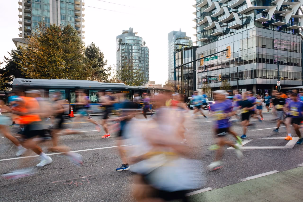 Motion-blurred runners race through a downtown Vancouver intersection during a marathon, passing in front of a TransLink bus as glass high-rise towers and city streets frame the background