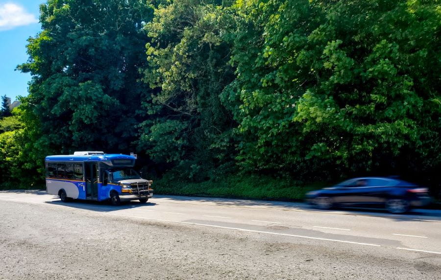 Community shuttle bus travelling down rural road with trees in background.